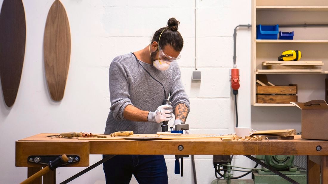 Man using a rotary tool to carve wood, with 2 wooden surfboards hanging on the wall in the background