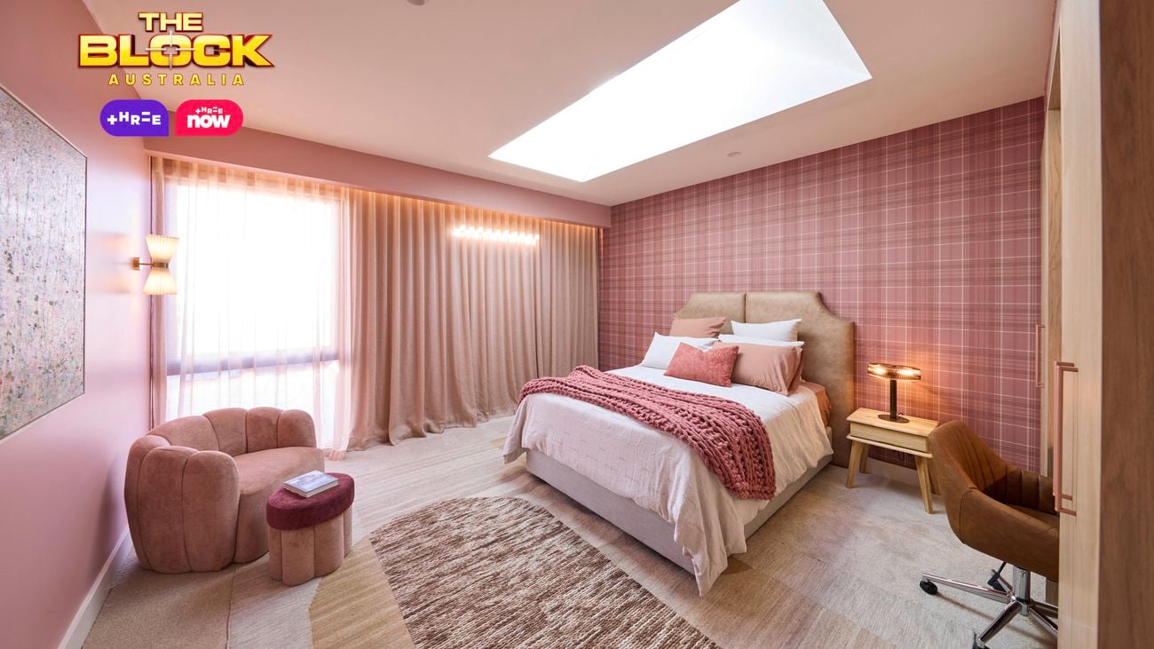 Pink bedroom with a check wallpapered wall and a double bed with pink linens. Opposite is a large velvet chair and matching foot stool.