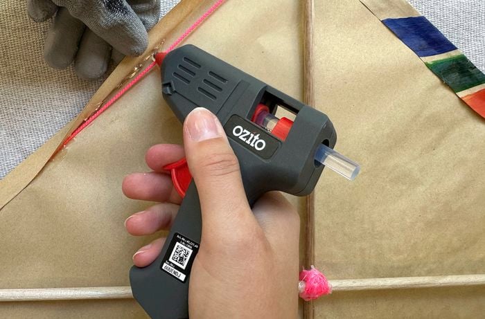 Hand using a hot glue gun to glue a pink string along the edge of a brown paper kite frame with wooden dowels.