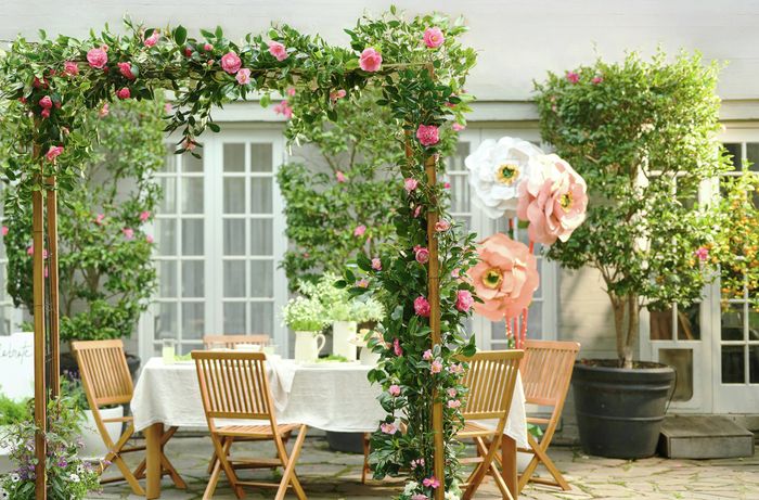 Outdoor garden arch with pink flowers and foliage in front of a dining table, with a paved patio and grassy lawn