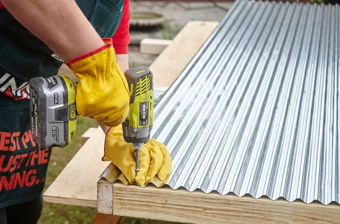 A person attaching a sheet of corrugated iron to a timber frame using a cordless drill