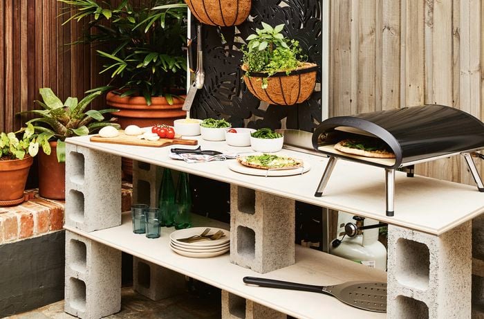 Two hands hold a wall basket holding a variety of herbs and hanging on a decorative outdoor screen.