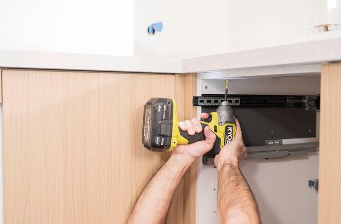 A person is using a cordless drill to add L brackets for extra support and and help lock the benchtop in place.