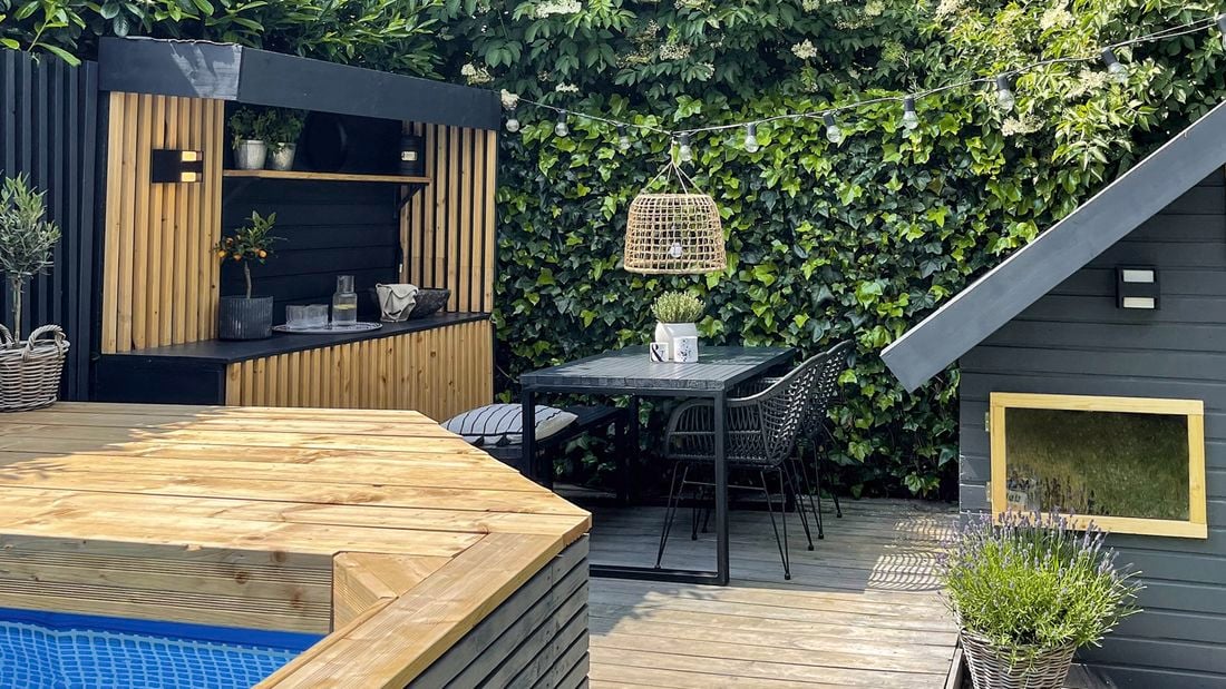 A timber deck with a table and chairs, built-in buffet and the edge of a timber-framed pool in the foreground and a shed to the right.