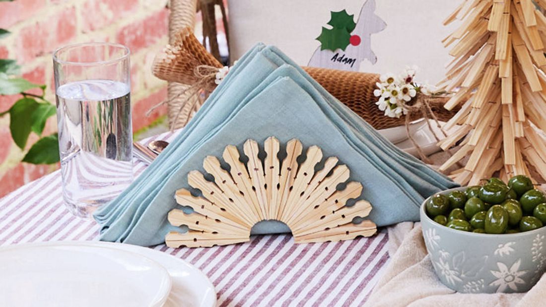 A semicircular napkin holder crafted from wooden pegs holding a pale blue folded napkin on a table setting.