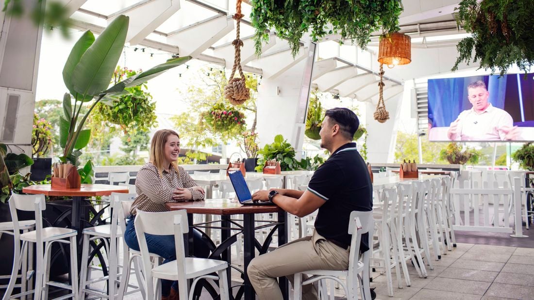  A man and a woman sit across from each other at a table in a bright, modern outdoor café area with white furniture and hanging plants. The man is working on a laptop while they converse. The setting includes lush greenery, decorative lighting, and a large screen in the background displaying a person speaking.