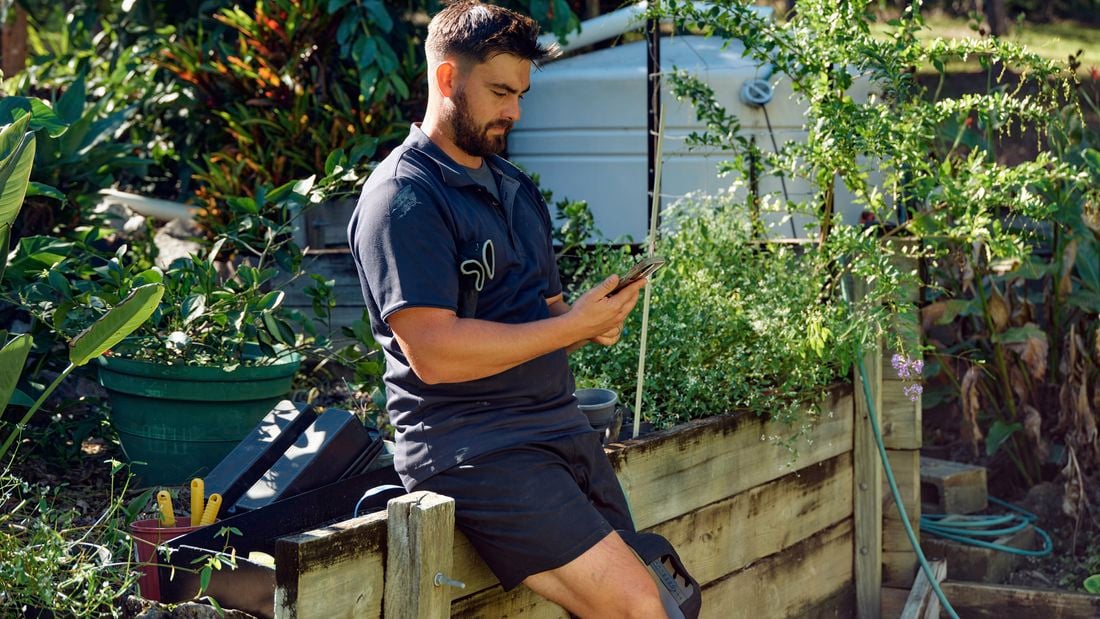 A landscaper sits on a timber retaining wall outside looking at his phone