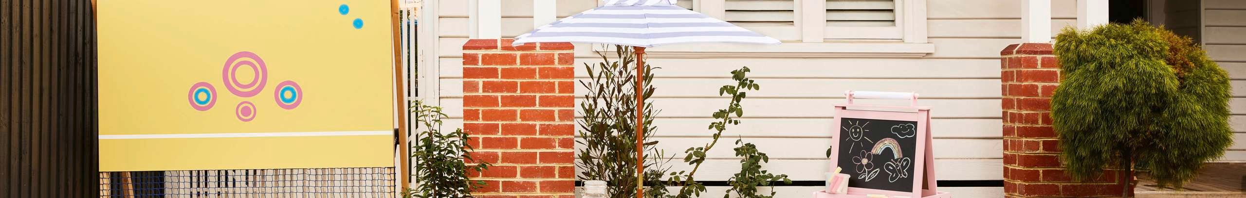 Children’s play area in a front yard with a tennis net, striped umbrella, chalkboard easel and garden plants in front of a weatherboard house.