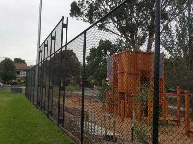 Fenced playground with orange climbing equipment next to a grassy field, with trees, houses, and parked cars in the background.