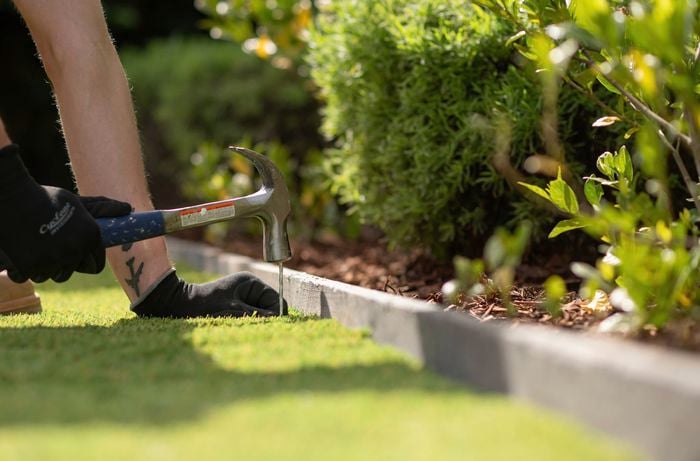 A person wearing gloves is using a hammer to secure the synthetic turf edge with turf pegs.