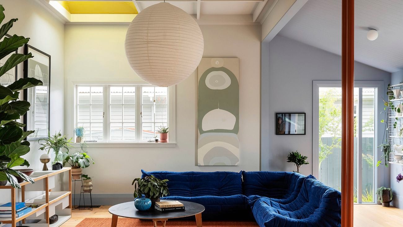  Living room with a deep blue sofa, white round paper lantern, low coffee table on a rust-coloured rug, and potted plants. White shuttered window and tall abstract artwork on the wall, with an opening to a grey-painted adjoining room