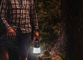 A person wearing a plaid shirt and backpack holding a lit camping lantern in a dark forest.