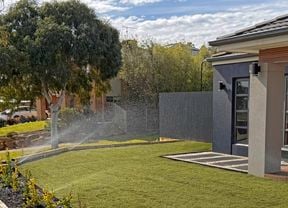 A front yard garden design with a smooth lawn being watered by pop up sprinklers in front of a modern grey house with a timber patio, and a pebbled and paved path.