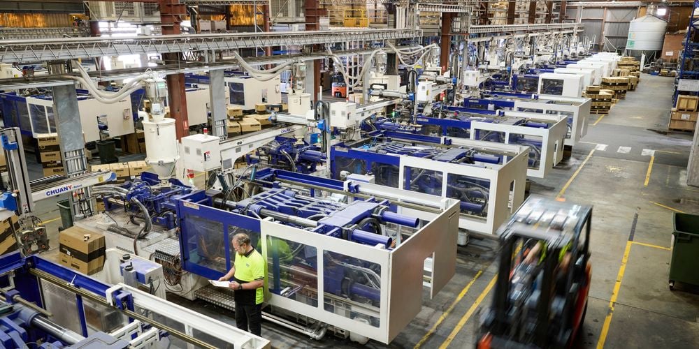 Wide-angle
  view of a busy manufacturing facility with multiple machines in operation. A
  worker in a hi-vis yellow shirt monitors one of the machines, while a
  forklift drives past in the aisle.