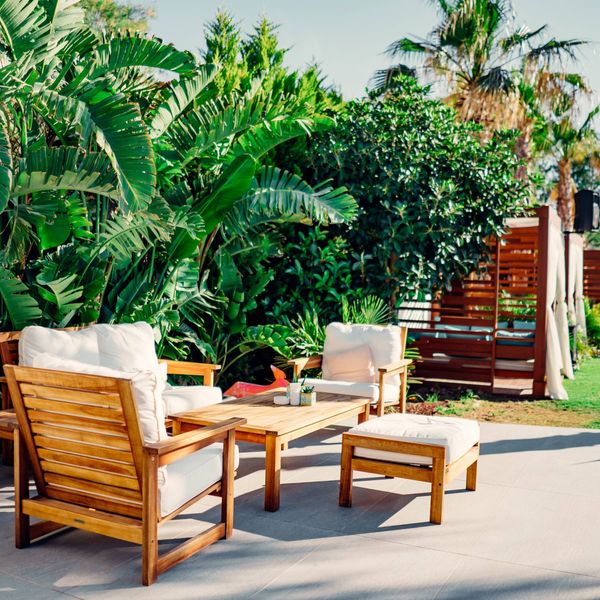 Outdoor patio with wooden lounge furniture and white cushions, surrounded by lush tropical plants and palm trees in a landscaped garden.