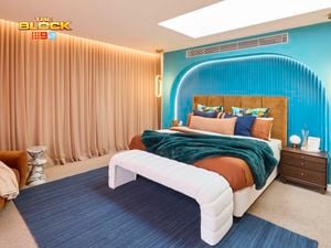A modern bedroom featuring a large bed with white and brown bedding, colorful pillows, and a green throw blanket. The blue headboard wall has an arched design and pendant lights on either side. A wooden nightstand with decorative items sits next to the bed. The room includes a blue rug, beige curtains, and a rectangular skylight in the ceiling. 