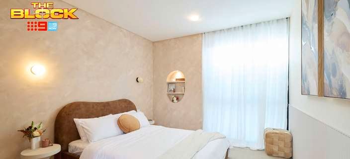 A furnished bedroom, with a bed with a side table next to it, with shelving and two paintings on the wall, carpet on the floor and The Block on the image.