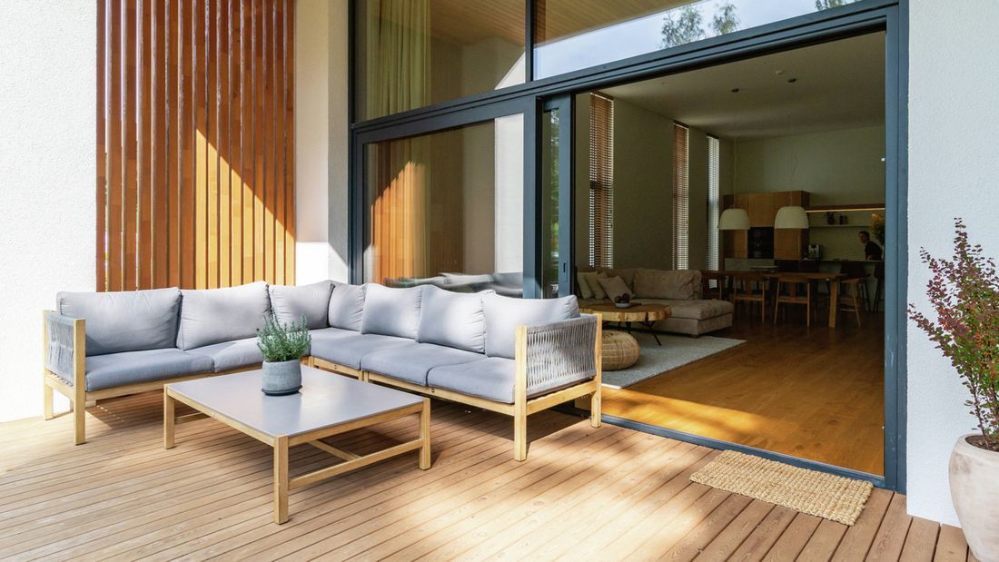Modern outdoor wooden deck with a grey sectional sofa and coffee table, looking into a bright interior space through large sliding glass doors.