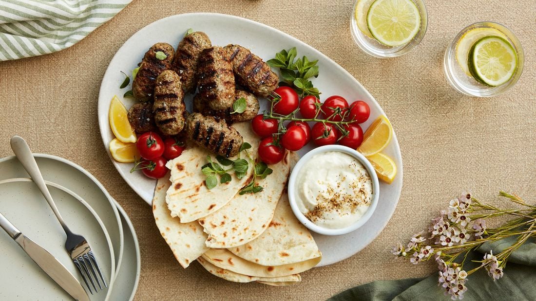 Lamb kofta on a white platter with pita bread, a bowl of creamy garlic sauce, cherry truss tomatoes and lemon wedges garnished with oregano.