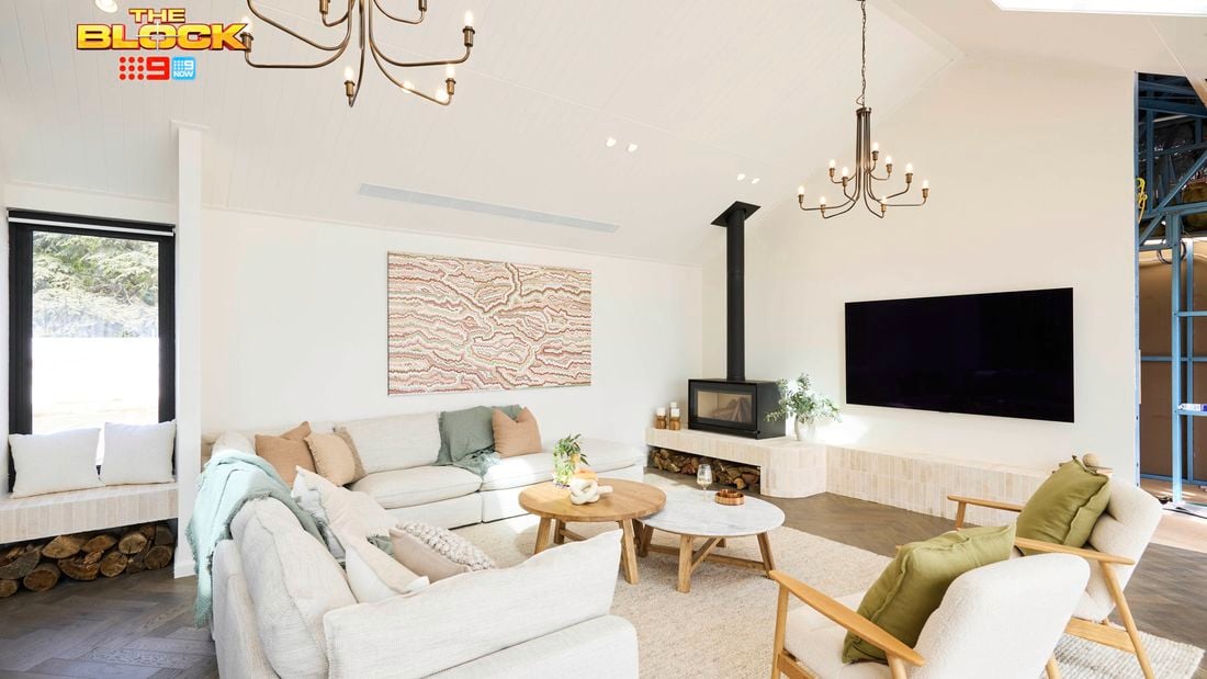 A modern living room with high ceilings, featuring two chandeliers, a wall-mounted flat-screen TV, and a fireplace with logs stored below. The space includes a light-colored sectional sofa with cushions and throws, two armchairs with green cushions, and two round wooden coffee tables. Abstract artwork hangs above the sofa, and large windows bring in natural light. Decorative plants are placed throughout. 