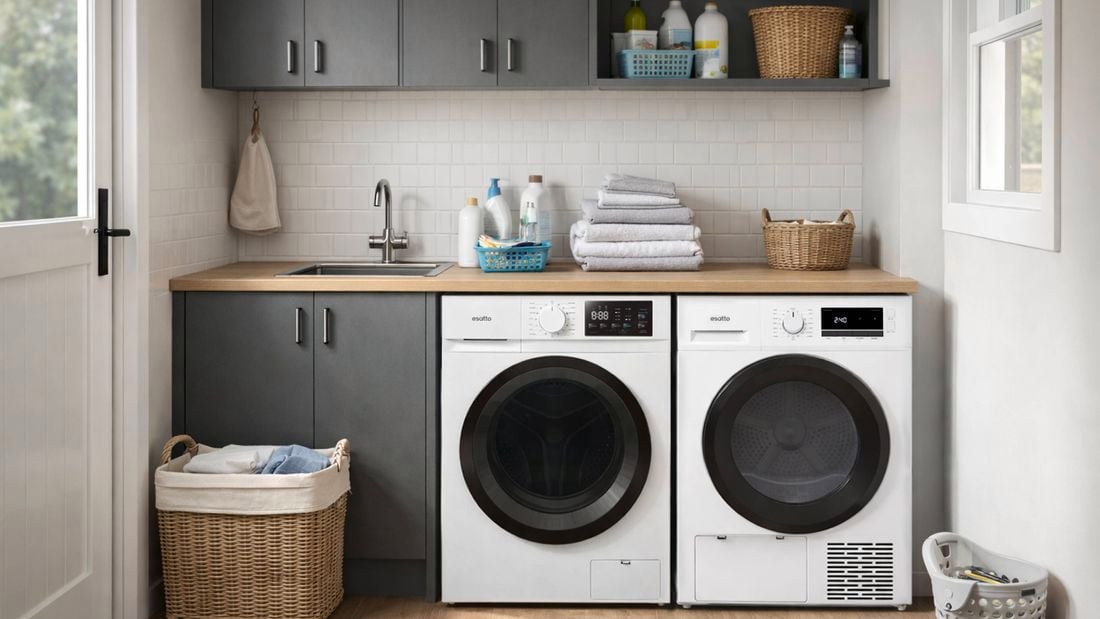Modern laundry with grey cabinetry, wooden benchtop, and two front‑loading machines side by side beneath a sink, featuring folded towels, cleaning bottles, wicker baskets, and a laundry hamper on timber flooring beside a window.