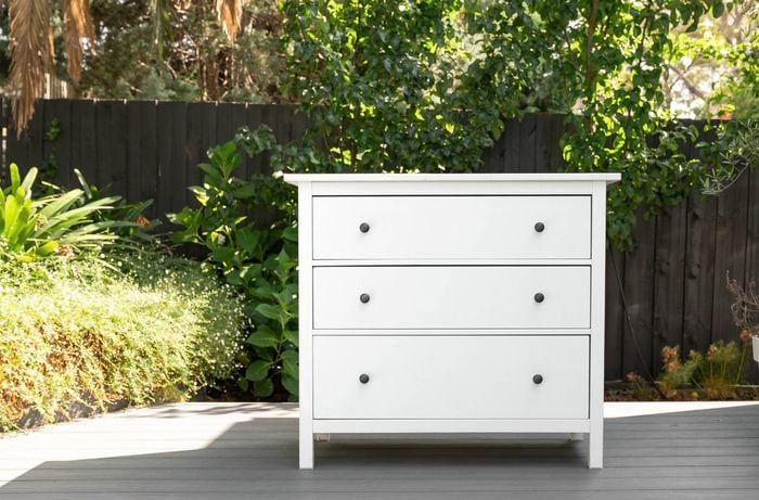 White three-drawer cabinet positioned on an outdoor deck with plants in the background.