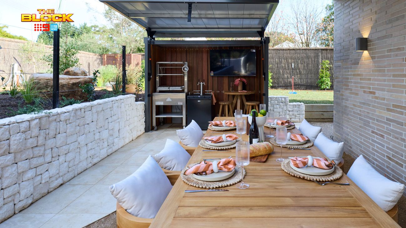 Covered outdoor dining area featuring a timber table set for dining, cushioned bench seating, stone walls and an outdoor kitchen.