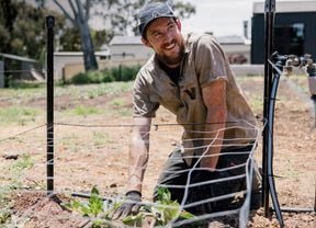 A tradesperson wearing a cap, work shirt, and gloves kneeling outdoors with a mesh fence in front of him.]