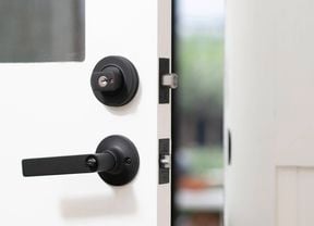 A white door with a black deadbolt lock and handle is shown partially open, revealing the extended latch.