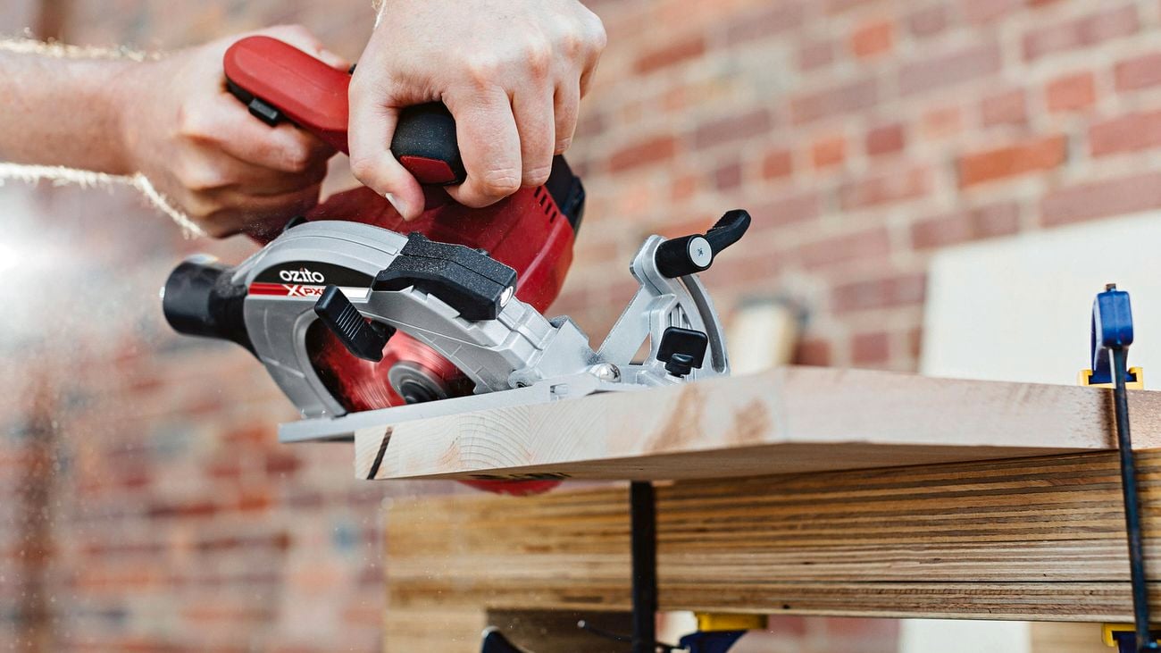 A person holding an Ozito circular saw at an angle against a piece of wood secured to a workbench.