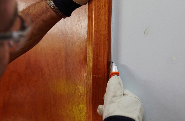 Person using a box cutter to create a gap between architrave and wall.