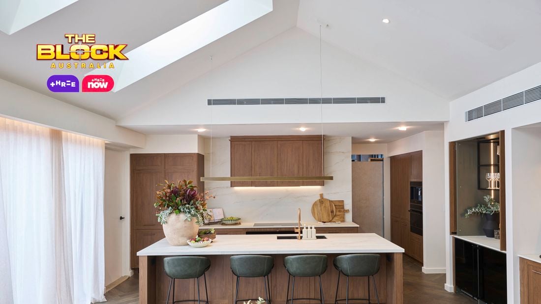 A modern white kitchen with brown cupboards, bar stools at the benchtop with a vase with flowers in it and two bowls with items in them on it.