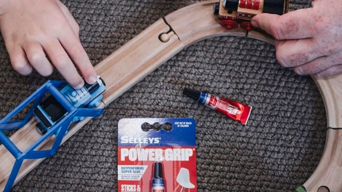 Close up of two hands directing two toy trains toward one another on a wooden toy track. A tube of Selleys Power Grip super glue is in centre