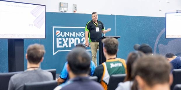 A presenter stands at a podium addressing an audience at a Bunnings Expo seminar, with attendees seated and looking toward the stage and display screens.