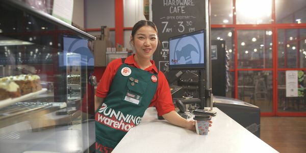 Hardware store café counter with a team member standing behind the service area holding a takeaway coffee cup. A glass display cabinet, point-of-sale screens and café signage are visible, with the seating area located in the background.