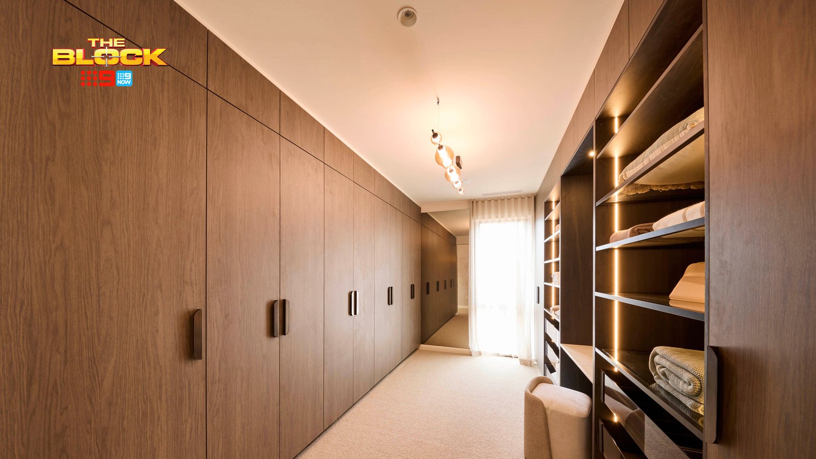 A well-organized walk-in closet featuring wooden cabinetry on both sides. The left side has closed cabinets with handles, while the right side displays open shelves with neatly folded clothes and towels. A small cushioned stool sits near the shelves on a carpeted floor. The space is illuminated by ceiling lights and natural light from a window at the far end. 