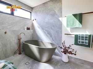 Modern bathroom with matte gray freestanding tub, stone-look gray tiles, wall-mounted faucet, green towels on wooden racks, and natural light from a high window. 