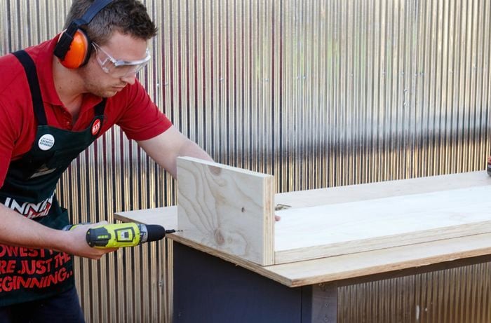 A person wearing protective gear drilling through two panels of plywood joined at right angles