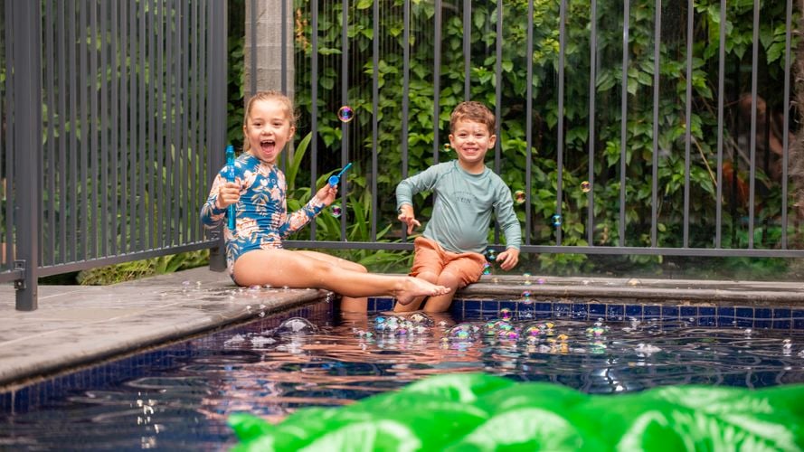 Two kids sitting on an edge of a pool with fencing surrounding the pool.