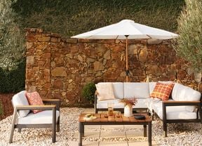Outdoor patio lounge setting featuring a white-cushioned sectional sofa and matching armchair around a wooden coffee table on white gravel. A large white umbrella shades the seating area, with a rustic stone wall, manicured hedge, and olive trees creating a landscaped garden backdrop. Decorative cushions, a vase of flowers, and drinks on the table add a warm, inviting atmosphere.