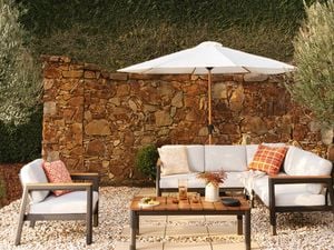 Outdoor patio lounge setting featuring a white-cushioned sectional sofa and matching armchair around a wooden coffee table on white gravel. A large white umbrella shades the seating area, with a rustic stone wall, manicured hedge, and olive trees creating a landscaped garden backdrop. Decorative cushions, a vase of flowers, and drinks on the table add a warm, inviting atmosphere.