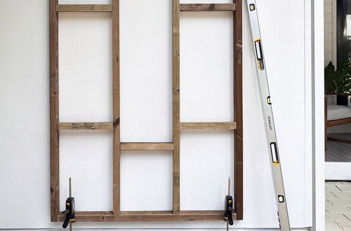A timber frame mounted against a white wall, with a spirit level leans on the right and two clamps holding the base.