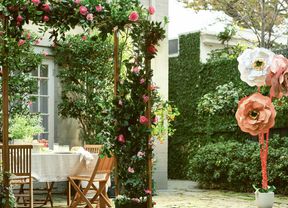 Outdoor garden arch with pink flowers and foliage in front of a dining table, with a paved patio and grassy lawn