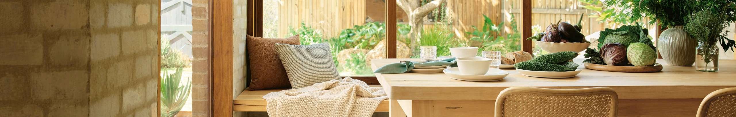 Sunlit dining and living area with a timber table set for a meal, bench seating by a window, indoor greenery, and warm natural textures.