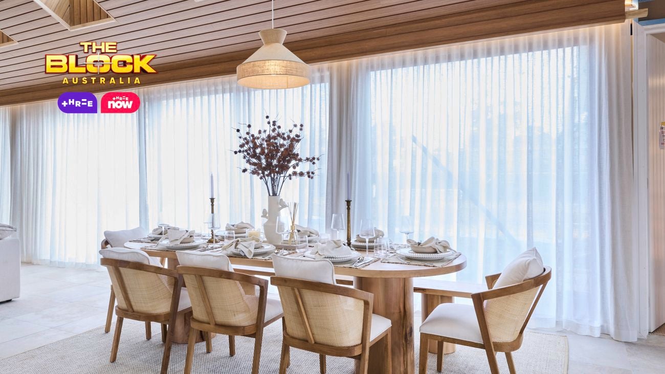 A dining room featuring a wooden table set for six, with plates, glasses, napkins, candles, and a central vase of flowers. The space is illuminated by natural light through large windows with sheer white curtains. A hanging light fixture is suspended from the wooden-paneled ceiling, and a light-colored rug lies beneath the dining set.