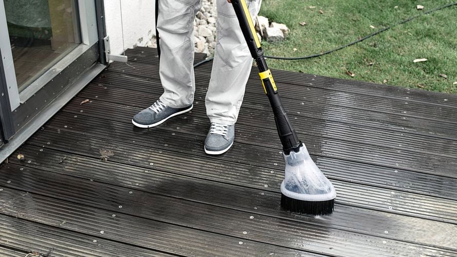 Person cleaning a wooden deck with a pressure washer and brush attachment, with grass and rocks in the background.