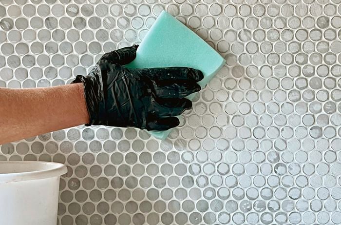 A person wearing a disposable glove removes excess grout from a newly tiled splashback with a damp sponge.