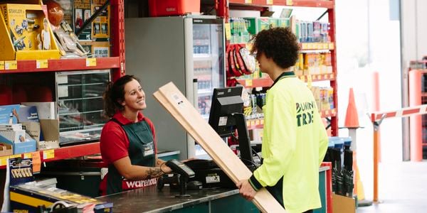 A Bunnings Trade specialist carrying lengths of timber talking to a Bunnings team member at a warehouse checkout