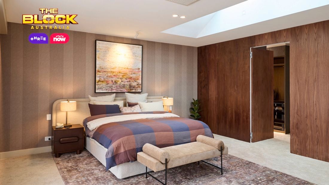 A spacious, modern bedroom with a large bed dressed in a plaid blanket and pillows. The room includes a skylight, a wooden accent wall, a built-in fireplace with a seating area, and large windows with curtains that allow natural light to enter.