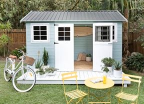 A painted timber garden shed with a bike and garden furniture in a backyard